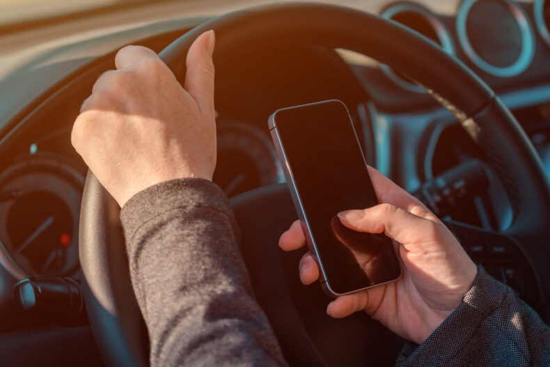 close up of a person's hand and who is distracted driving by operating a car and text messaging on a cell phone with one hand holding smartphone and other hand holding the steering wheel