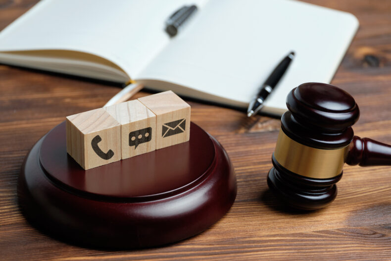 wooden blocks with a phone symbol, text symbol, and email symbol on top of a gavel plate, near a brown gavel, and open notebook on a desk, to symbolize contact a local personal injury attorney